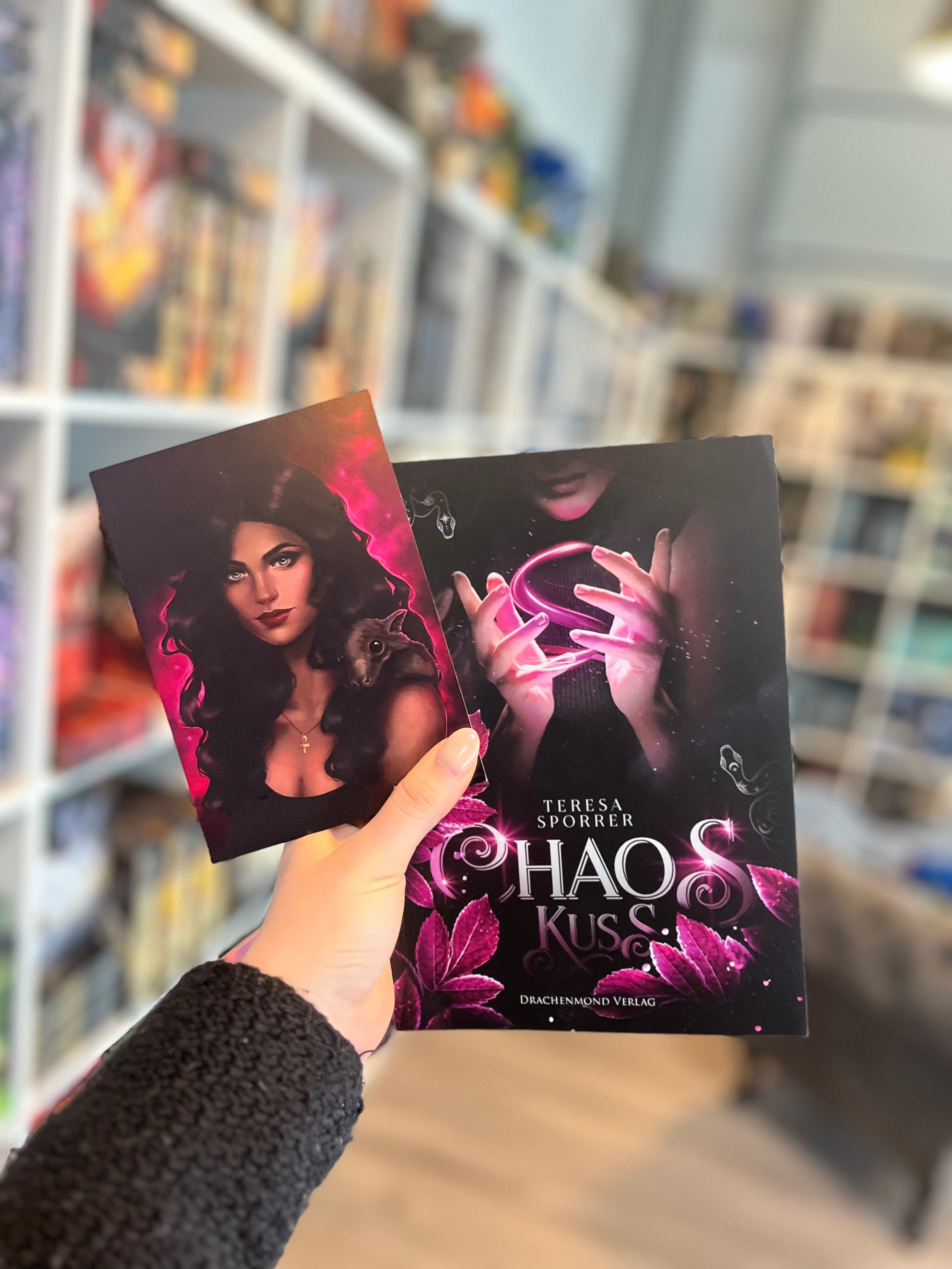 Das Bild zeigt das Buch "Chaoskuss" und die zugehörige Charakterkarte. Auf der Karte ist die Protagonistin May vor einem pinken Hintergrund abgebildet. Auf ihrer Schulter sitzt eine Fledermaus.