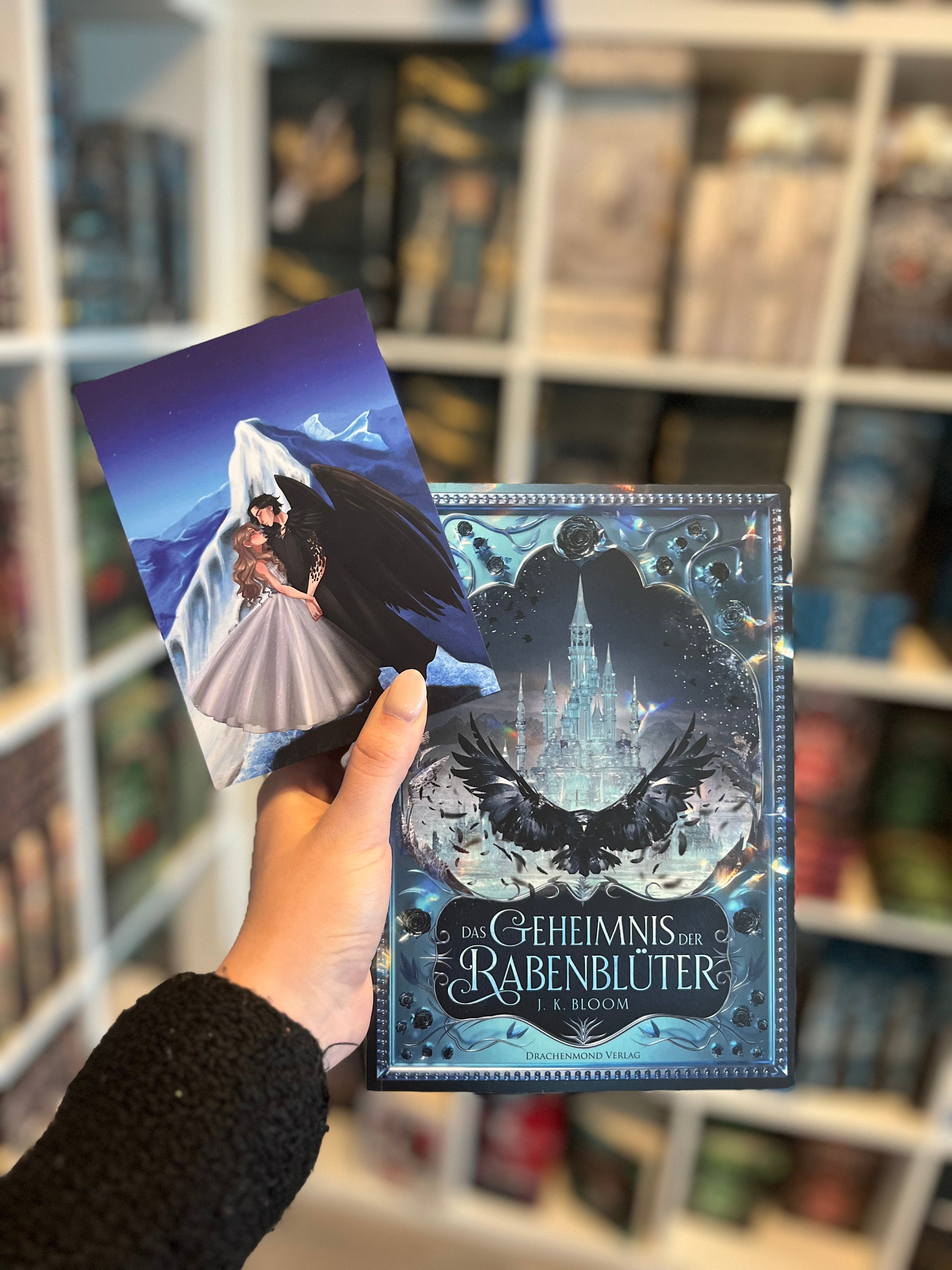 Auf dem Bild sieht man eine Hand, welche das Buch "Das Geheimnis des Rabenblüter" und die dazugehörige Charakterkarte hält. Auf der Karte sieht man die Protagonistin und den Protagonisten, wie sie gemeinsam vor einem Berg stehen. Der Protagonist trägt ein schwarzes Gewand und hat schwarze Flügel auf dem Rücken. Die Protagonistin trägt ein silbernes Kleid.