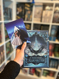 Auf dem Bild sieht man eine Hand, welche das Buch "Das Geheimnis des Rabenblüter" und die dazugehörige Charakterkarte hält. Auf der Karte sieht man die Protagonistin und den Protagonisten, wie sie gemeinsam vor einem Berg stehen. Der Protagonist trägt ein schwarzes Gewand und hat schwarze Flügel auf dem Rücken. Die Protagonistin trägt ein silbernes Kleid.