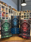 Das Bild zeigt drei Stapel von "Die 12 Häuser der Magie" - Band 1 bis 3 in Blau, Rot und Grün. Das Cover ist dabei direkt auf den Betrachter gerichtet und der Farbschnitt sowie der Buchrücken bei den liegenden Büchern sichtbar.
