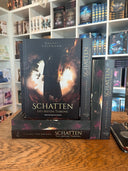 Das Bild zeigt einen Stapel von "Schatten des roten Throns". Das Cover ist dabei direkt auf den Betrachter gerichtet und der Farbschnitt sowie der Buchrücken bei den liegenden Büchern sichtbar.