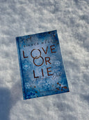 Das Bild zeigt das Buch "Love or Lie" von vorn. Das Cover ist dabei direkt auf den Betrachter gerichtet; es zeigt ein hellblaues Schneelockenmuster mit goldenen Sprenkeln.