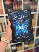 Das dunkelblaue Cover zeigt einen großen mit Blumen verzierten Rahmen. In der Mitte ist ein großer Schmetterling, der in Blau-, Weiß- und Silbertönen leuchtet. Darüber steht in großen Buchstaben der Titel, darunter die Autorin.