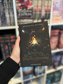 Das dunkle Cover zeigt mittig eine Phiole, in der ein Feuer brennt. Dahinter ist ein großer schwarzer Drachenkopf mit leuchtenden goldenen Augen. Der Titel steht im unteren Bildbereich in goldenen Buchstaben. Auf dem Farbschnitt sind Flammen und ein goldenes Drachenauge abgebildet.