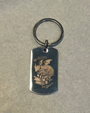 Das Produktbild zeigt einen rechteckigen Schlüsselanhänger (mit abgerundeten Ecken) auf beigen Grund. Auf ihm ist das Bild eines Drachen, der auf einem Bücherstapel sitzt, sowie der Schriftzug "Im Herzen ein Drache" zu sehen.