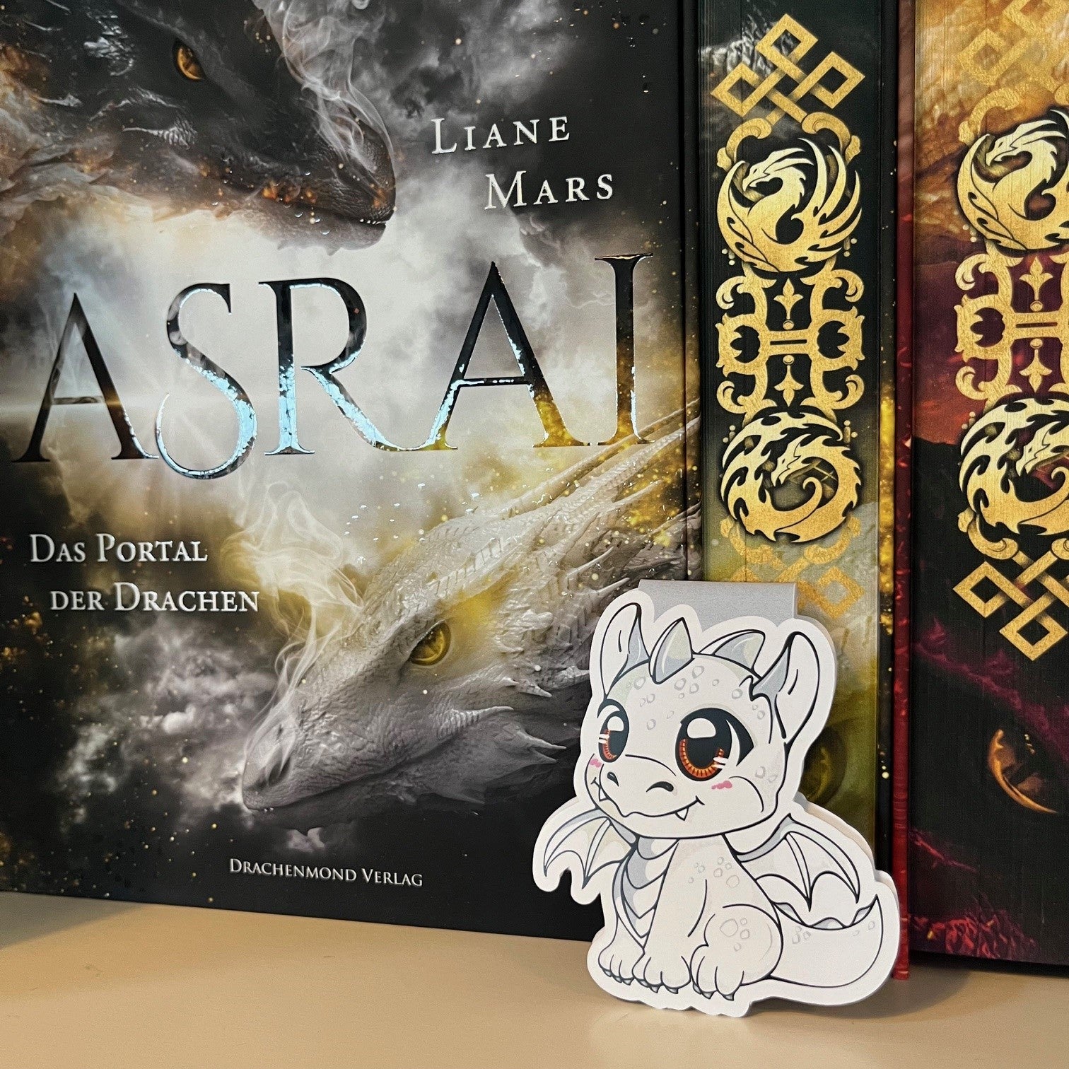 Das Produktbild zeigt ein Magnetlesezeichen mit einem weißen Babydrachen. Im Hintergrund ist das Buch 