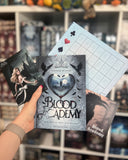 Auf dem Bild sieht man eine Hand, welche das Buch "Blood Academy" hält. Zu sehen sind drei Zusatzkarten zu dem Buch. Links ist eine Charakterkarte, die eine junge, blonde Frau zeigt. Sie steht vor einem dunklen Schloss. Rechts sieht man eine Karte mit einem Raben. Oberhalb des Buches sieht man eine Art Bingo-Karte, welche zu dem Buch gehört.