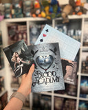 Auf dem Bild sieht man eine Hand, welche das Buch "Blood Academy" hält. Zu sehen sind drei Zusatzkarten zu dem Buch. Links ist eine Charakterkarte, die eine junge, blonde Frau zeigt. Sie steht vor einem dunklen Schloss. Rechts sieht man eine Karte mit einem Raben. Oberhalb des Buches sieht man eine Art Bingo-Karte, welche zu dem Buch gehört.