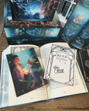 Auf dem Bild liegen die Charakterkarte und der Signiersticker auf dem aufgeschlagenen Buch "Book Elements".  Die Charakterkarte zeigt die Protagonistin Lin und den Feuerelementar Ric, die sich gegenüberstehen und einander in die Augen schauen. In Rics linker Hand lodert eine Flamme, um Lin wirbelt eine Windhose. Der Signiersticker ist mit dem Rahmen vom Buchcover verziert.