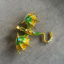 Das Produktbild zeigt eine Brosche in Form eines Drachen, mit ausgebreiteten Flügeln. Auf dem Rücken und dem Schwanz sind jeweils drei helle runde Edelsteine, auf den Flügeln jeweils ein grüner ovaler Edelstein. Der Drache ist grün und golden.