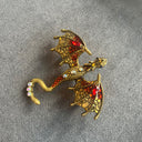 Das Produktbild zeigt eine Brosche in Form eines Drachen, mit ausgebreiteten Flügeln. Auf dem Rücken und dem Schwanz sind jeweils drei helle runde Edelsteine, auf den Flügeln jeweils ein roter ovaler Edelstein. Der Drache ist rot und golden.