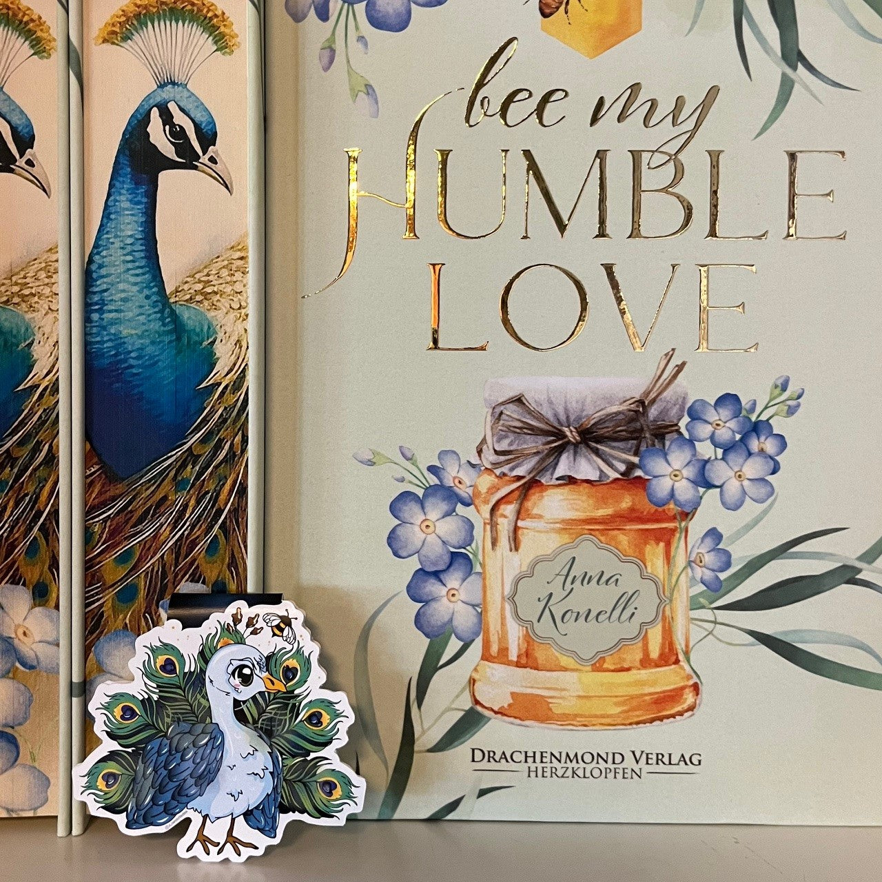 Das Produktbild zeigt ein Magnetlesezeichen mit einem Pfau. Oben links fliegt eine kleine Hummel.