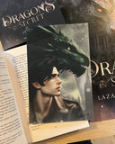Auf dem Produktbild sieht man das Page Overlay des Hauptcharakters Caleb im Profil vor dem Profil eines grünen Drachen. Es liegt auf einem geöffneten Buch, daneben sieht man noch den Teil eines Coverposters und darüber das Covers des Buches. 