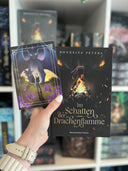 Das Bild zeigt, wie eine Hand das Buch "Im Schatten der Drachenflamme" von Roukeiya Peters und die Charakterkarte mit der Hauptfigur vor den Drachen des Buches hält. 