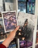 Auf dem Bild ist eine Hand abgebildet, welche das Buch "Magie aus Tod und Kupfer" und die dazugehörige Charakterkarte hält. Auf der Karte sind die beiden Protagonisten in Kampfstellung abgebildet. Im Hintergrund sieht man eine große Motte und einen Torbogen mit verschnörkelten Verzierungen.