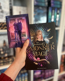 Auf dem Bild ist eine Hand abgebildet, welche das Buch "Monstermagie" und die dazugehörige Charakterkarte hält. Auf der Karte sind die Protagonistin und der Protagonist abgebildet, welche Arm in Arm vor einem Schrank stehen.
