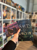 Auf dem Bild sieht man eine Hand, welche das Buch "Salt & Sorcery" und die dazugehörige Charakterkarte hält. Auf der Karte sieht man die Protagonistin und den Protagonisten, welche gemeinsam auf einer Wiese sitzen.