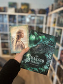 Auf dem Bild sieht man eine Hand, welche das Buch "Der Smaragddrache" und die dazu gehörende Charakterkarte hält. Auf der Charakterkarte sieht man die Protagonistin Gemma im Seitenprofil. Ihre blonden Haare sind nach hinten geflochten und flattern leicht im Wind.