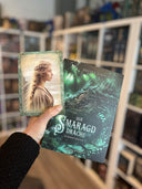 Auf dem Bild sieht man eine Hand, welche das Buch "Der Smaragddrache" und die dazu gehörende Charakterkarte hält. Auf der Charakterkarte sieht man die Protagonistin Gemma im Seitenprofil. Ihre blonden Haare sind nach hinten geflochten und flattern leicht im Wind.