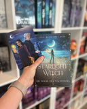 Auf dem Bild sieht man eine Hand, welche das Buch "Starlight Witch" als Softcover mit der dazu passenden Charakterkarte hält. Auf der Charakterkarte sind die Protagonistin und der Protagonist abgebildet.