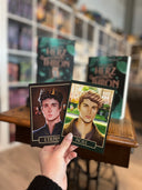 Auf dem Bild sieht man eine Hand, welche die beiden Charakterkarten zu "Zwischen Herz und Thron" hält. Auf der linken Karte sieht man das Portrait eines jungen Mannes mit braunen Haaren. Unter ihm steht der Name "Etienne". Auf der rechten Karte sieht man das Portrait eines blonden Mannes. Unter ihm steht der Name "Noel".