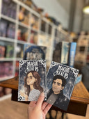 Auf dem Bild sieht man im Hintergrund das Buch "Magiestaub" und im Vordergrund hält eine Hand die dazugehörenden Charakterkarten. Die linke Karte zeigt die Protagonistin mit braunen Haaren und einem silbernen Oberteil. Auf der rechten Karte sieht man den Protagonisten mit braunen Haaren und einem dunklen Mantel.