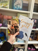 Auf dem Bild sieht man eine Hand, welche das Buch "Sommergold" und die dazu gehörende Charakterkarte hält. Auf der Karte sieht man die Protagonistin und den Protagonisten, welche zusammen in einer Hängematte sitzen und etwas trinken.