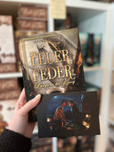 Das Bild zeigt, wie eine Hand das Buch "Feuerfeder - Briefe an den König" von Anne Danck und (davor) die Charakterkarte mit den beiden Hauptfiguren des Buches hält. 