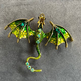 Das Produktbild zeigt eine Brosche in Form eines Drachen, mit ausgebreiteten Flügeln. Auf dem Rücken und dem Schwanz sind jeweils drei helle runde Edelsteine, auf den Flügeln jeweils ein grüner ovaler Edelstein. Der Drache ist grün und golden.