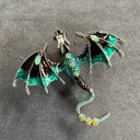 Das Produktbild zeigt eine Brosche in Form eines Drachen, mit ausgebreiteten Flügeln. Auf dem Rücken und dem Schwanz sind jeweils drei helle runde Edelsteine, auf den Flügeln jeweils ein türkiser ovaler Edelstein. Der Drache ist türkis, schwarz und silber.