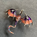 Das Produktbild zeigt eine Brosche in Form eines Drachen, mit ausgebreiteten Flügeln. Auf dem Rücken und auf dem Schwanz sind jeweils drei runde Edelsteine, auf den Flügeln jeweils ein heller ovaler Edelstein. Der Drache ist lila und rosa.