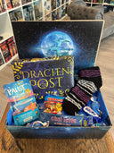 Das Produktbild zeigt vorn eine geöffnete Drachenpost in blauem Schuppen-Design. Man sieht im Deckel der Box das Verlagslogo - einen Drachen, der vor dem Mond fliegt - und in der Box die zum Thema Drachentraum passenden Goodies: Kuschelsocken, Badezusatz, Gute-Nacht-Tee und andere Kleinigkeiten. Im Hintergrund sieht man die Bücherregale des Verlagsladens. 