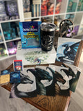 Auf dem Produktbild sieht man den Inhalt der Drachenbox #drachenliebe: einen schwarzen Tumbler, fünf Page Overlays mit Drachen, Gummibärchen, Tee und einen Keks. Im Hintergrund sieht man die Bücherregale im Drachennest.