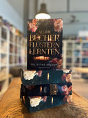 Das Bild zeigt einen Stapel von "Als die Bücher flüstern lernten". Das Cover ist dabei direkt auf den Betrachter gerichtet und der Farbschnitt sowie der Buchrücken bei den liegenden Büchern sichtbar.