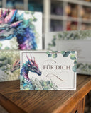Das Bild zeigt eine Karte mit einem Drachenkopf in Lila- und Grüntönen, umgeben von Eukalyptus und dem Schriftzug "Für Dich". Im Hintergrund sieht man verschwommen die dazugehörige Buchbox.