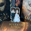 Das Produktbild zeigt ein Magnetlesezeichen mit einer Frau in einem silberweißen Ballkleid und ein Mann in einem schwarzen Anzug mit grüner Krawatte. Sie hält in ihrer Hand eine Silberne Maske, er  hält in seiner Hand eine schwarze Maske. Gezeichnet ist das Lesezeichen im Anime-Stil.