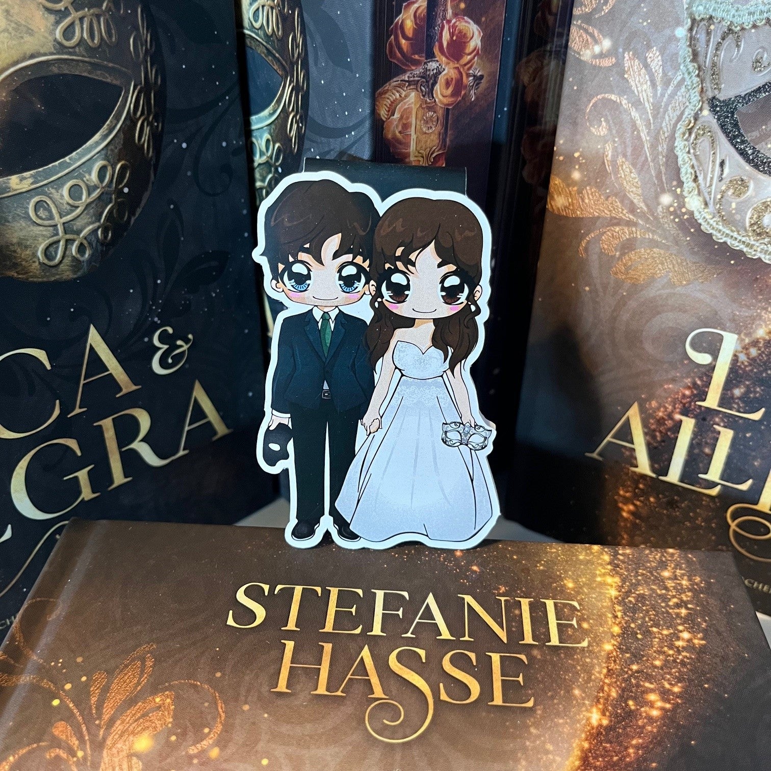 Das Produktbild zeigt ein Magnetlesezeichen mit einer Frau in einem silberweißen Ballkleid und ein Mann in einem schwarzen Anzug mit grüner Krawatte. Sie hält in ihrer Hand eine Silberne Maske, er  hält in seiner Hand eine schwarze Maske. Gezeichnet ist das Lesezeichen im Anime-Stil.