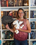 Das Bild zeigt die Autorin Liane Mars, welche vor einem Bücherregal steht und einen weißen Stoffdrachen auf dem Arm hat. In der rechten Hand hält sie ein Schild, auf dem "Bekommt den Drachen" steht.