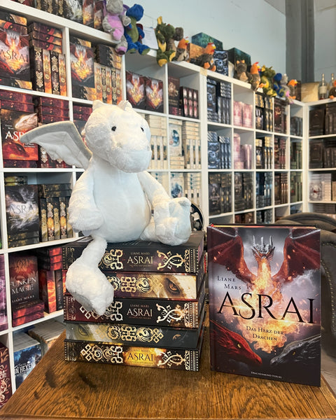 Das Produktbild zeigt den weißen Plüsch-Drachen Manila (aus der Asrai-Buchreihe von Liane Mars) von vorn auf einem Stapel Asrai-Bücher sitzend vor Bücherregalen. Daneben sieht man frontal das Buch 