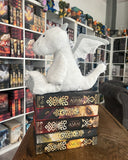 Das Produktbild zeigt den weißen Plüsch-Drachen Manila (aus der Asrai-Buchreihe von Liane Mars) von der Seite auf einem Stapel Asrai-Bücher sitzend vor Bücherregalen. 
