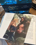 Das Bild zeigt das Page Overlay zum Buch "Die Legenden der Unaschkin" von Lena Klassen auf einem aufgeschlagenen Buch liegend. Auf dem Page Overlay befinden sich die beiden einander zugewandten Hauptfiguren der Geschichte in einem Dschungel. 