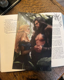 Das Bild zeigt das Page Overlay zum Buch "Die Legenden der Unaschkin" von Lena Klassen auf einem aufgeschlagenen Buch liegend. Auf dem Page Overlay befinden sich die beiden einander zugewandten Hauptfiguren der Geschichte in einem Dschungel. 