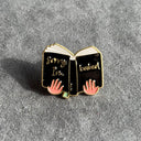 Das Produktbild zeigt einen Pin in Form eines aufgeschlagenen Buches, gehalten von zwei Händen. Das Buch ist schwarz mit einem grünen Lesebändchen. Auf dem linken Einband steht "Sorry I'm" und auf dem rechten Einband steht "booked".