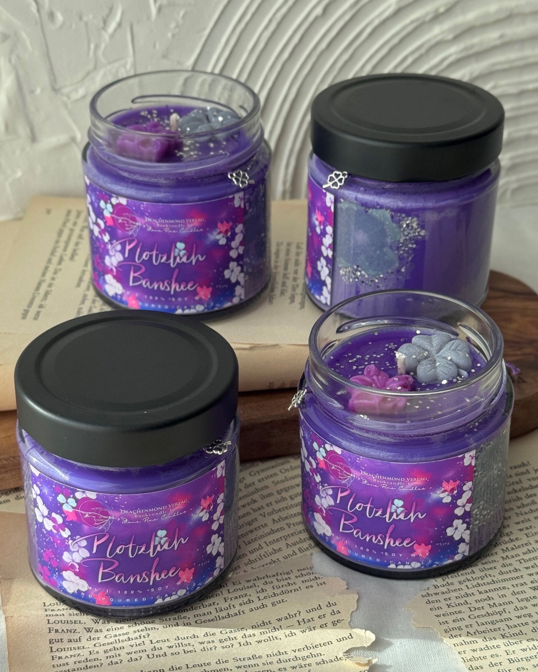 Das Produktbild zeigt zwei Kerzen im Glas. Die linke Kerze ist mit einem schwarzen Deckel verschlossen. Darunter hängt ein lilafarbenes Band mit einem Glücksklee-Anhänger. Auf dem lilafarbenen Label steht mittig der Titel in weiß-rosa. Darum herum sind weiße, lilafarbene und rosa Blüten verteilt. Die rechte Kerze ist geöffnet. Neben dem Docht sind zwei Blüten aus Wachs, eine rosa und eine grau. Der Wachs der Kerze ist lila mit silbernem Glitter.