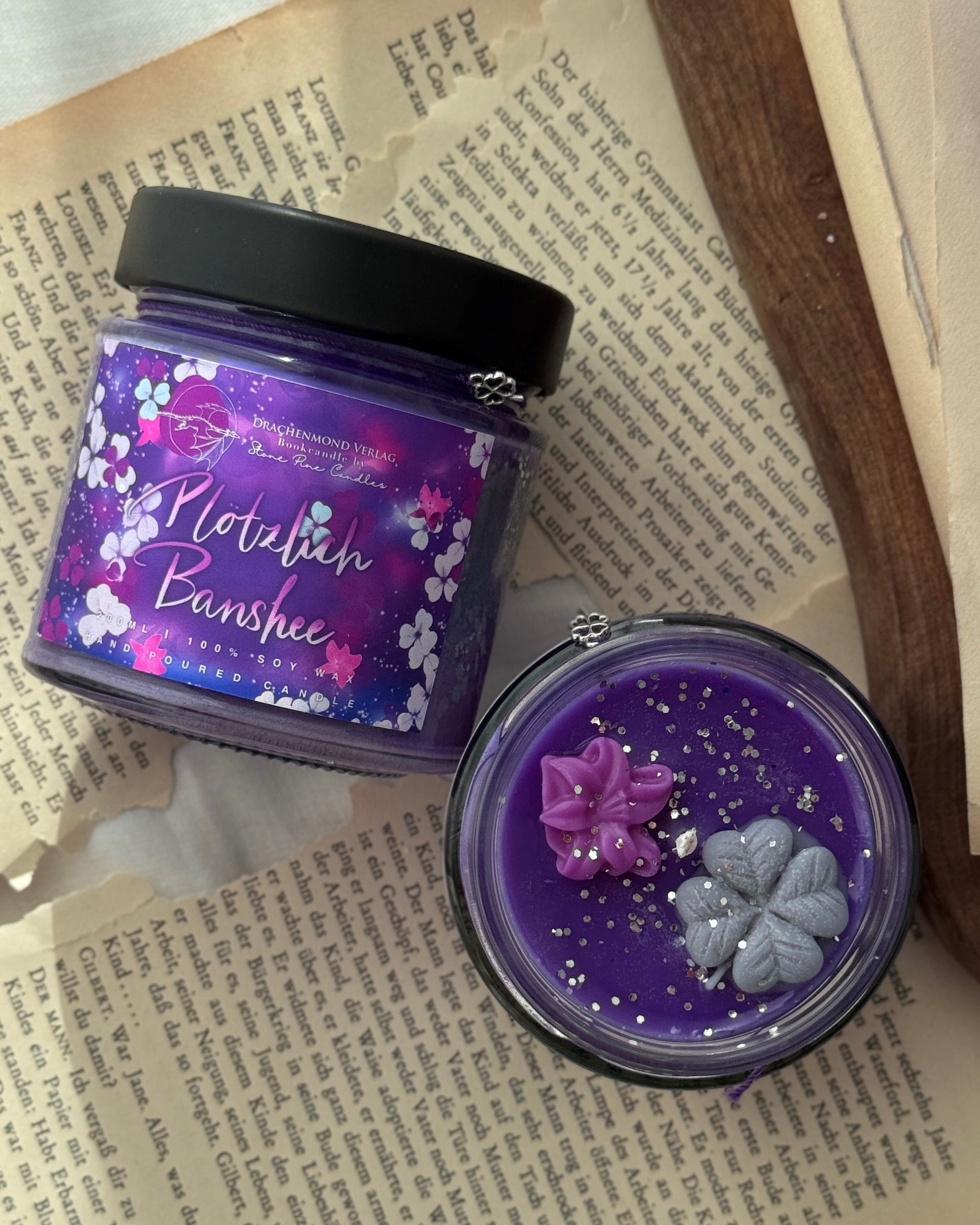 Das Produktbild zeigt zwei Kerzen im Glas. Die linke Kerze ist mit einem schwarzen Deckel verschlossen. Darunter hängt ein lilafarbenes Band mit einem Glücksklee-Anhänger. Auf dem lilafarbenen Label steht mittig der Titel in weiß-rosa. Darum herum sind weiße, lilafarbene und rosa Blüten verteilt. Die rechte Kerze ist geöffnet. Neben dem Docht sind zwei Blüten aus Wachs, eine rosa und eine grau. Der Wachs der Kerze ist lila mit silbernem Glitter.