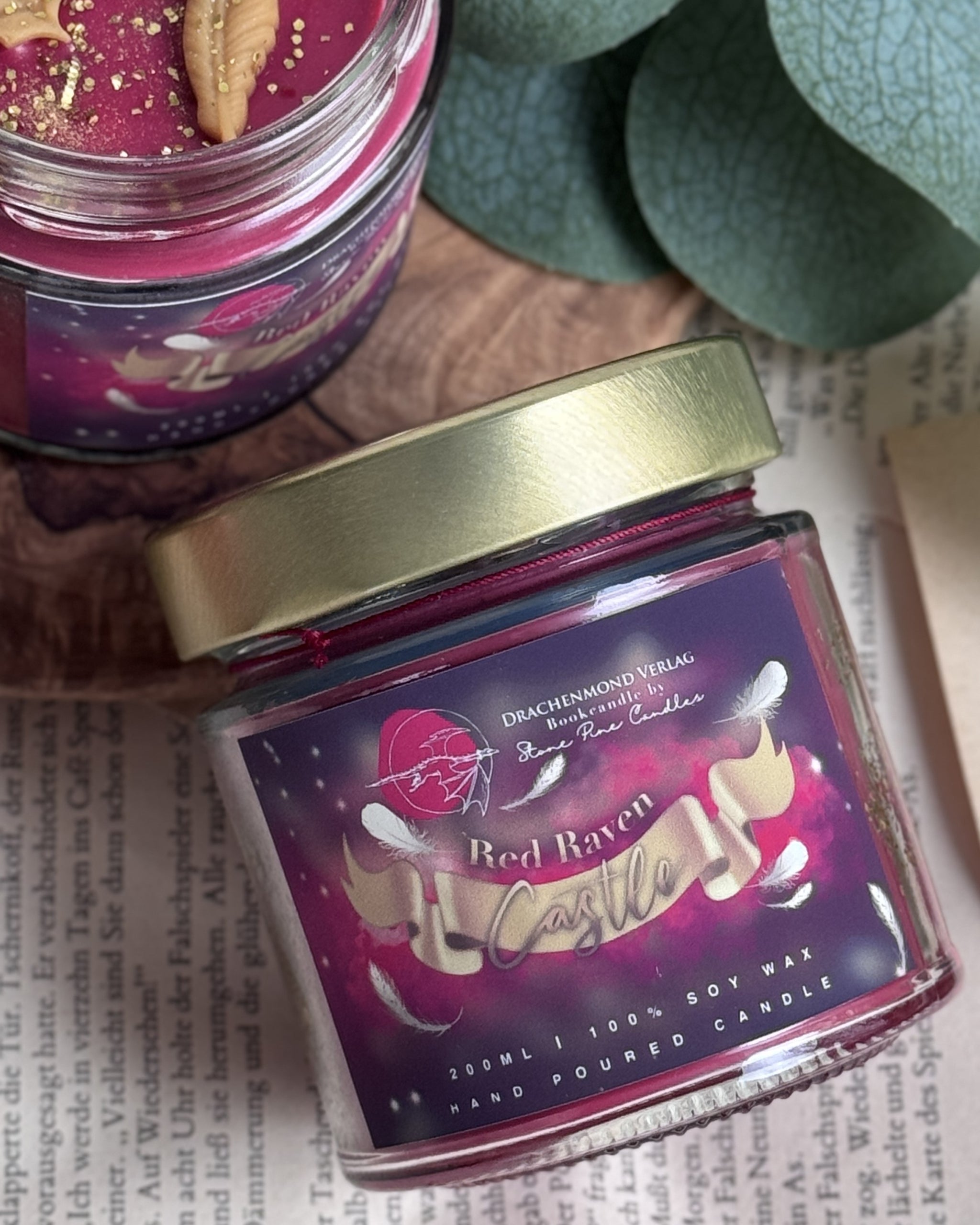 Das Produktbild zeigt eine liegende Kerze in einem Schraubglas mit goldenem Deckel. Auf dem Label steht mittig Red Raven und darunter ist eine goldene Banderole auf der das Wort Castle steht. Der Farbverlauf ist von außen lila nach innen zu rot.  