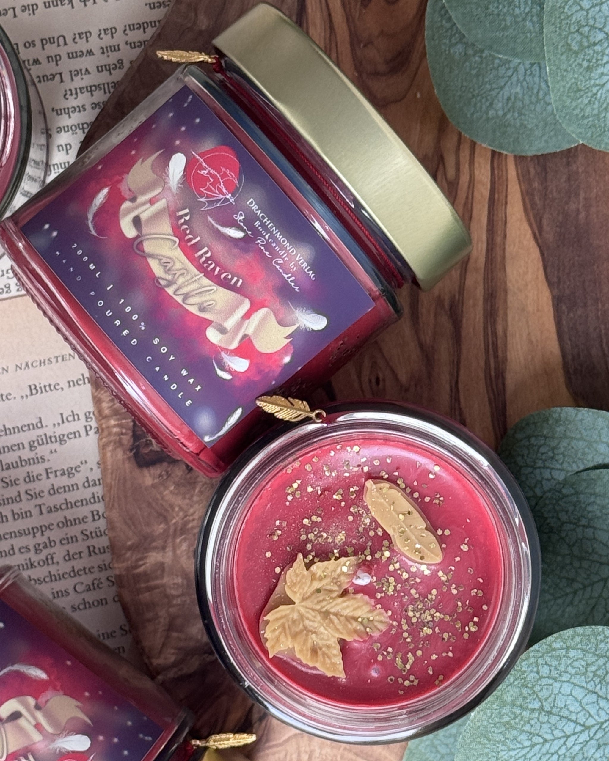 Das Produktbild zeigt eine Kerze im Glas von oben. Der Wachs ist rot mit goldenem Glitter, darauf befinden sich ebenfalls aus Wachs zwei unterschiedliche Blätter, neben dem Docht. Um das Glas herum hängt ein rotes  Band mit einem kleinen goldenen Anhänger in Form einer Feder.