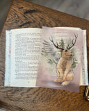 Auf dem Bild ist eine aufgeklappte Ausgabe von "Rosenrot" zu sehen. Auf den Seiten liegt ein Page Overlay, welches einen gezeichneten Wolpertinger zeigt. Das Buch liegt auf einer dunkelbraunen Unterlage mit Holzmaserung.