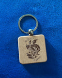 Das Produktbild zeigt einen quadratischen Schlüsselanhänger aus Holz. Auf ihm ist das Bild eines Drachen, der auf einem Bücherstapel sitzt, sowie der Schriftzug "Im Herzen ein Drache" zu sehen.