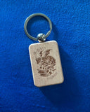 Das Produktbild zeigt einen rechteckigen Schlüsselanhänger aus Holz. Auf ihm ist das Bild eines Drachen, der auf einem Bücherstapel sitzt, sowie der Schriftzug "Im Herzen ein Drache" zu sehen.